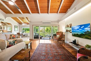 A spacious living room with a large rug, wooden ceiling beams, a sofa, a dining area, and a TV on the wall. - Island Home with AC - Close to Beaches and Shops (Princeville)
