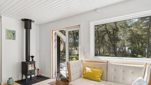 Een kamer met een bank, een raam met uitzicht op bomen, een open haard en een deur naar buiten.