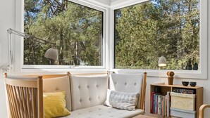 Een gezellige kamer met een bank, een klein tafeltje en een boekenkast. Grote ramen bieden uitzicht op bomen.