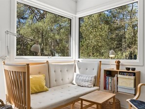 Ein gemütliches Zimmer mit einer Couch, einem kleinen Tisch und einem Bücherregal. Große Fenster bieten Blick auf Bäume.