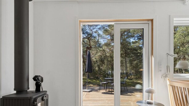 Een kamer met een houtkachel, een schuifdeur naar een terras buiten met een tafel en stoelen, en een kleine ronde tafel met een lamp.
