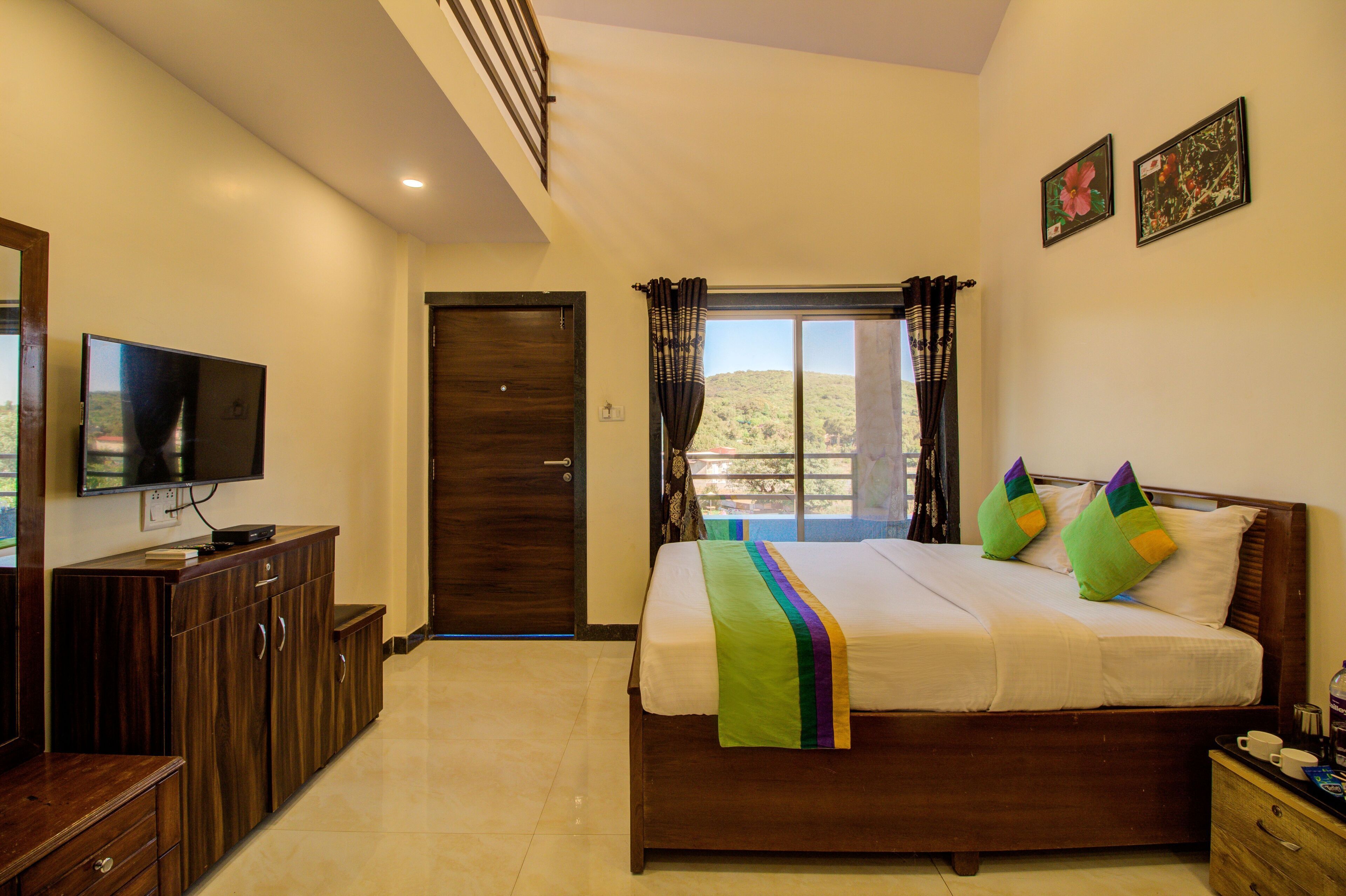 A bedroom with a bed, a TV, a wooden dresser, a window with curtains, and a door.