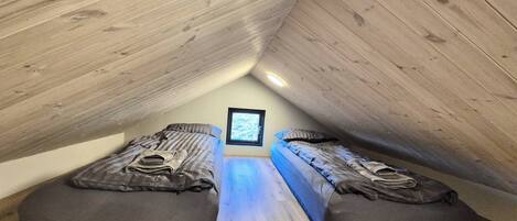 Una habitación pequeña con techo inclinado, dos camas, una ventana y suelo de madera.