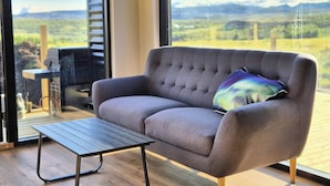 A modern living room with a grey sofa, a small wooden table, and a large window offering a view of a mountainous landscape. - Northern Lights Cabin - Syðribrú (Selfoss)