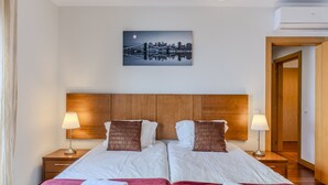 Habitación de hotel con dos camas, un cuadro de paisaje urbano, cabecera de madera y un equipo de aire acondicionado.