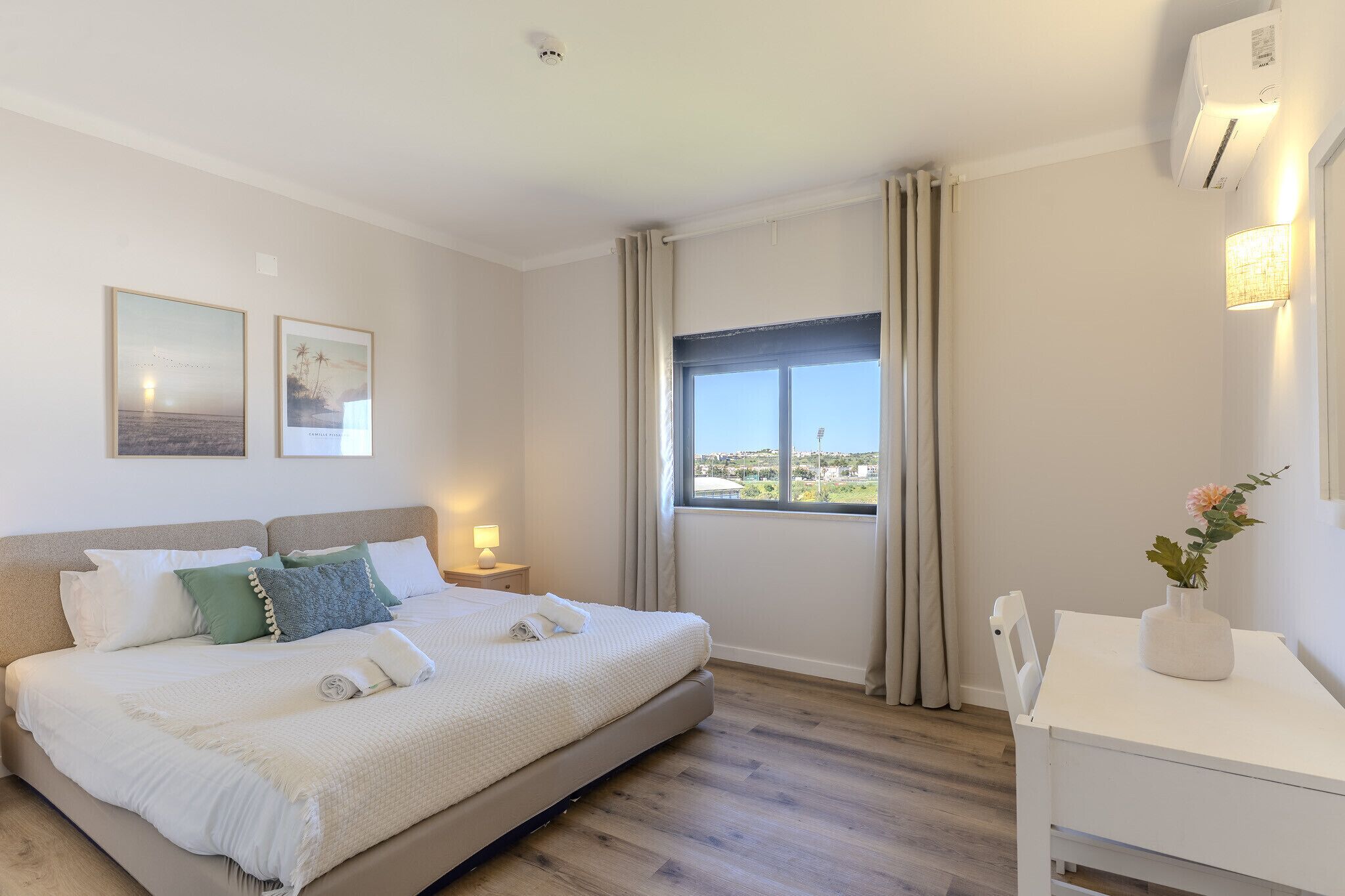 Une chambre à coucher comprenant un lit, une table de chevet, un bureau, une fenêtre avec des rideaux et un climatiseur fixé au mur.