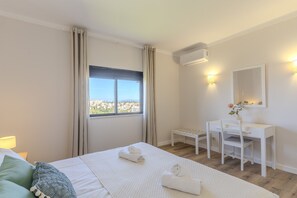 A hotel room with a bed, a desk, a chair, a window with curtains, and a view of buildings. - #319 Cozy 1 Bedroom Apartment | Pool & Relaxation (Albufeira)