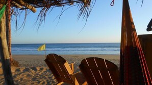 A beach scene with two wooden chairs under a thatched roof, a yellow flag, and the ocean in the background. - Casa Acalli Ocean View (San Pedro Pochutla)
