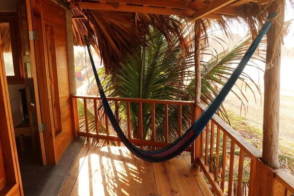 A wooden balcony with a hammock, wooden railings, and a view of palm trees.