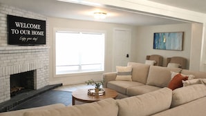 A living room with a white brick fireplace, a large window, a beige sectional sofa, and a 'WELCOME TO OUR HOME' sign. - Cozy Steinhatchee Getaway, Beachy + River Close (Steinhatchee)