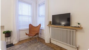 A room with a TV on a wooden shelf, a radiator, a chair, and a plant. - Charming Victorian Garden Apartment | Private Kitchen & Bathroom | King Bed (Southampton)