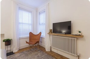 Ein Zimmer mit Fernseher auf einer Holzablage, einem Heizkörper, einem Sessel und einer Pflanze.