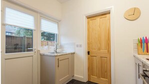 A kitchen with a wooden door, white cabinets, a window with blinds, and a clock on the wall. - Charming Victorian Garden Apartment | Private Kitchen & Bathroom | King Bed (Southampton)
