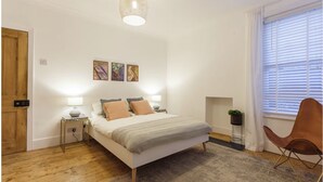 A bedroom with a bed, bedside lamps, a wooden floor, a window with blinds, and a wall-mounted artwork. - Charming Victorian Garden Apartment | Private Kitchen & Bathroom | King Bed (Southampton)