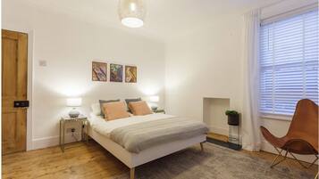 A bedroom with a bed, bedside lamps, a wooden floor, a window with blinds, and a wall-mounted artwork.