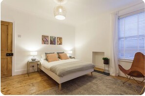 Ein Schlafzimmer mit einem Bett, Nachttischlampen, Holzboden, einem Fenster mit Jalousien und einem Wandbild.