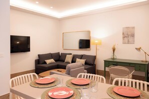 A modern living room with a sofa, a dining table set for four, a TV mounted on the wall, and a desk with a lamp.
