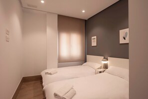 A hotel room with two beds, a nightstand with a lamp, a wall-mounted artwork, and a window with blinds.
