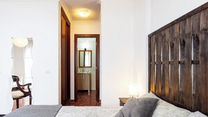 Une chambre à coucher avec un lit, une tête de lit en bois, une table de chevet avec une lampe, et une vue sur la salle de bain à travers une porte ouverte.