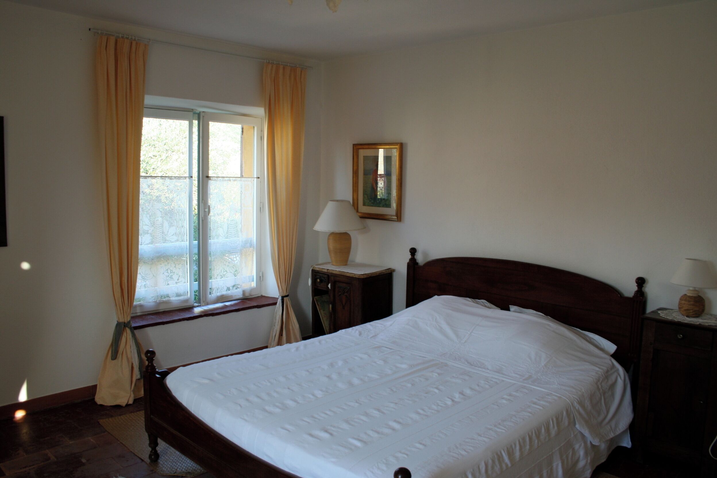 Een slaapkamer met een bed, een raam met gordijnen, een nachtkastje met een lamp en een schilderij aan de muur.
