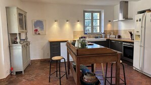 A kitchen with a wooden table, stools, a window, and modern appliances. - Near Cannes, authentic Bastide set in a garden planted with century-old olive trees (mouans sartoux)