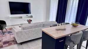 Una sala de estar moderna con televisor de pantalla plana, sofá gris, mesa de comedor con sillas y una mesita auxiliar con un florero.