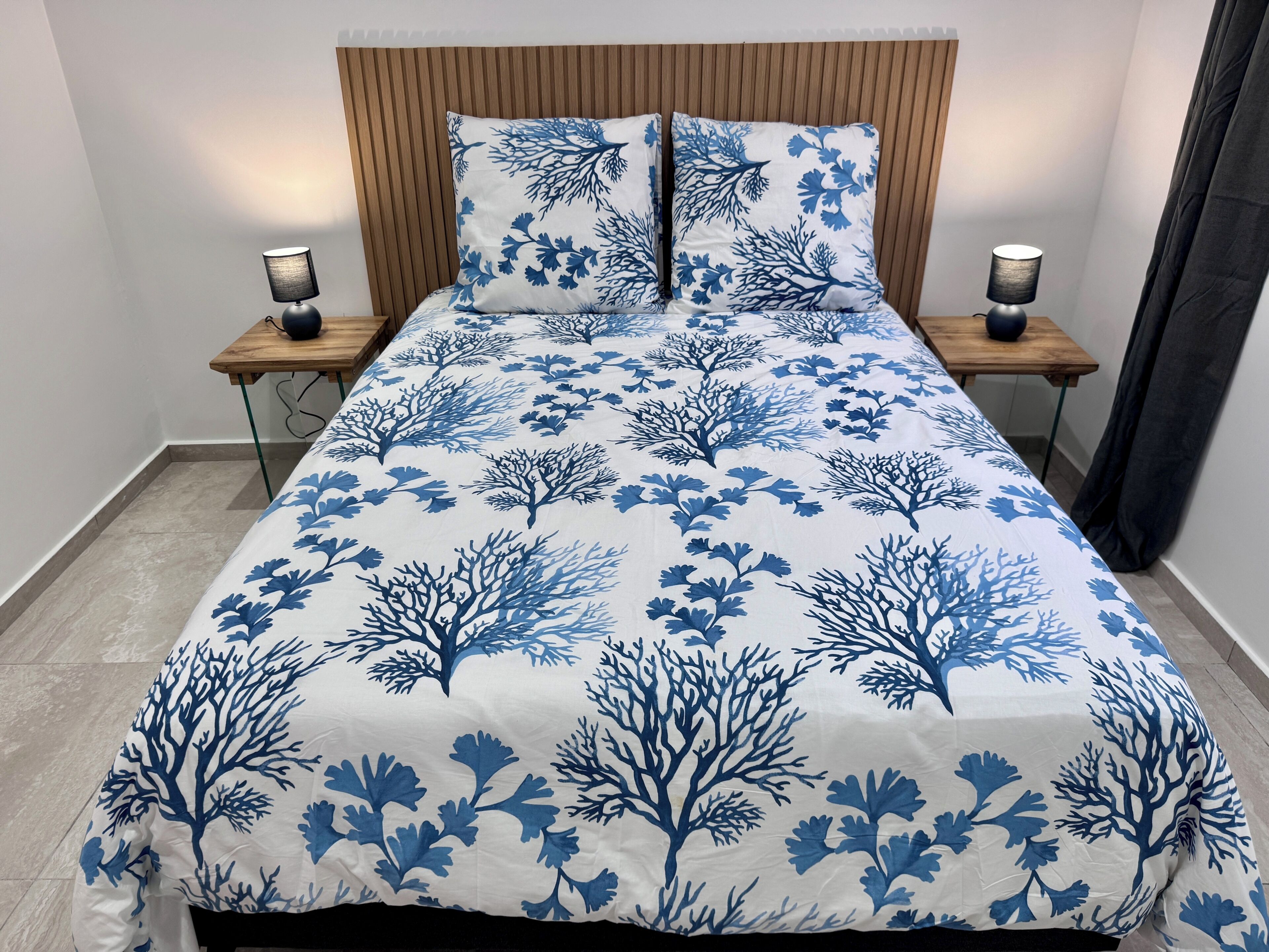 A bed with a blue and white coral pattern bedspread, two bedside lamps, and a wooden headboard.