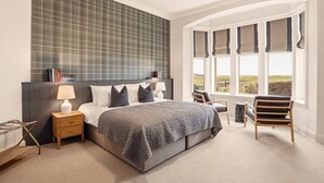 Ein Schlafzimmer mit Bett, Nachttischen, einem Sessel, einem Fenster mit Vorhängen und Blick ins Freie.