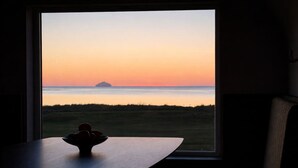 Ein Zimmer mit großem Fenster mit Blick auf ein Wiesfeld und ein Gewässer bei Sonnenuntergang.
