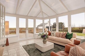 A spacious conservatory with large windows, a cushioned sofa, a coffee table, and a lamp. - Blackrock House – Iconic Home at Royal Troon (Troon)