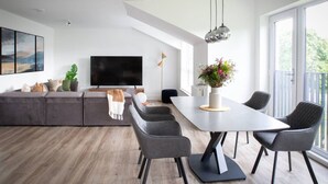 Ein modernes Wohnzimmer mit großem Flachbildfernseher, grauem Sofa, Esstisch mit Stühlen und einem Balkon mit Blick.