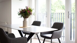 Ein Essbereich mit einem weißen Tisch, vier schwarzen Stühlen, einer Blumenvase und Blick auf Grün durch große Fenster.