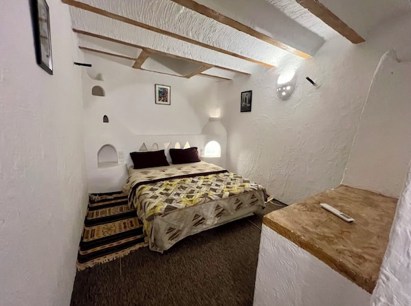 A bedroom with a bed, a rug, a wooden bench, and a wall with a window. - Gite TARIST (Bounoura)