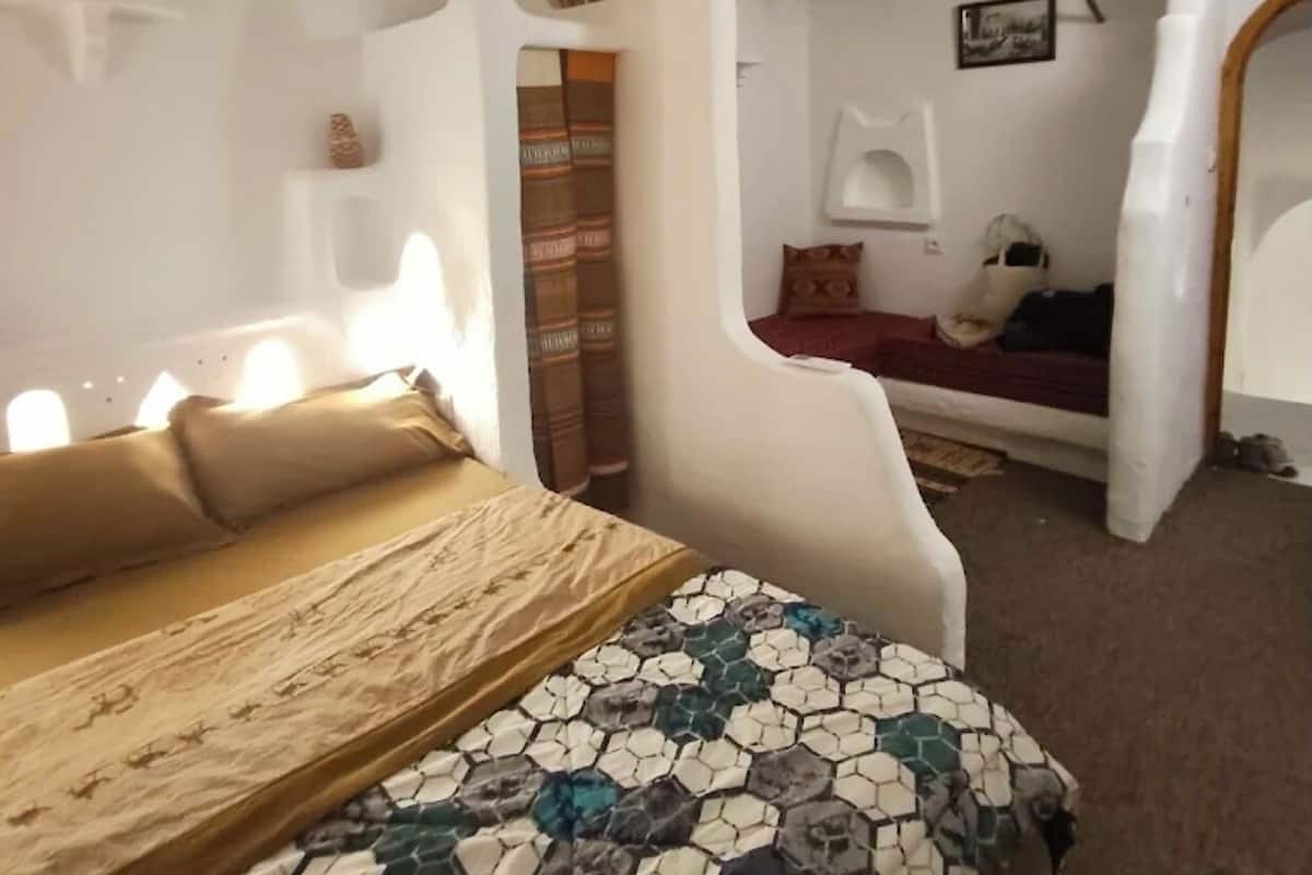A room with a bed, a chair, a small table, and a wall-mounted shelf.
