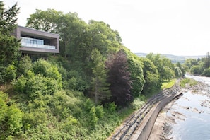 A modern building with a balcony overlooking a river and dense greenery. - Design-led Apartment | Central Pitlochry (Pitlochry)