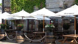 Outdoor tables with white umbrellas labeled 'Old Mill,' wooden barrels, and a stone building with ivy. - Design-led Apartment | Central Pitlochry (Pitlochry)