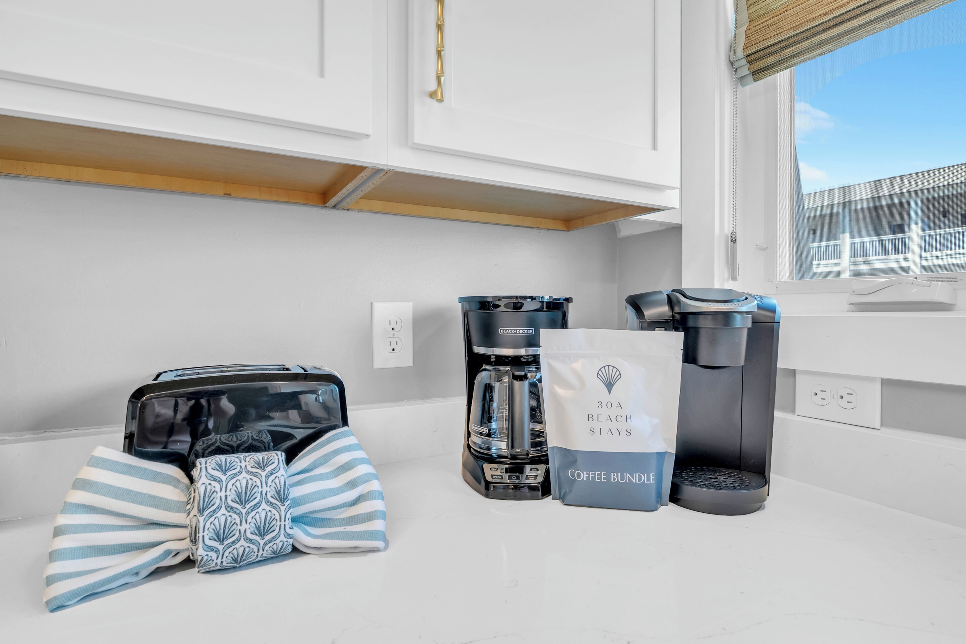 A kitchen with a coffee maker, a bag labeled '30A Beach Stays Coffee Bundle,' and a striped towel.