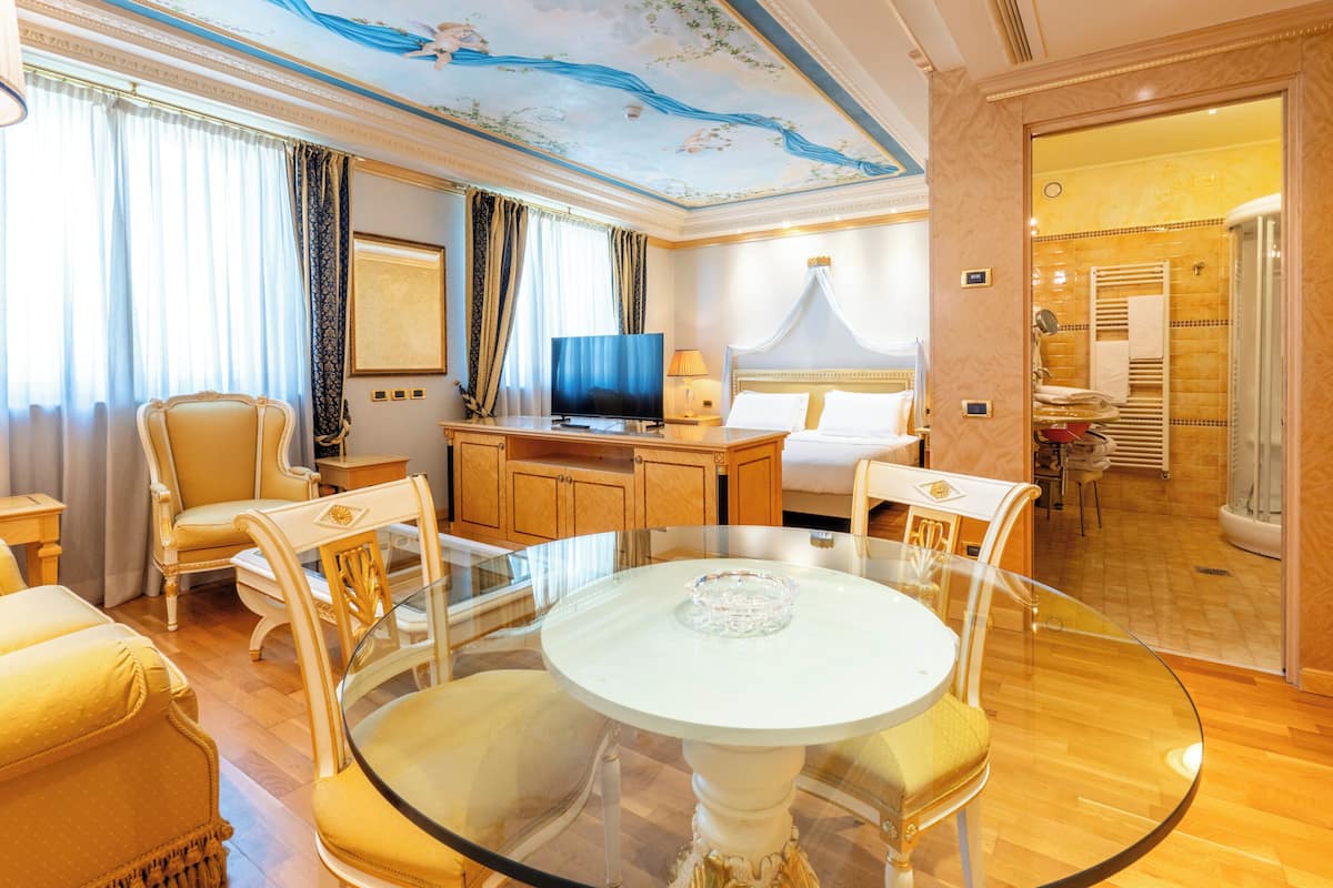 A hotel room with a glass dining table, a TV, a bed with a canopy, and a bathroom with a shower.