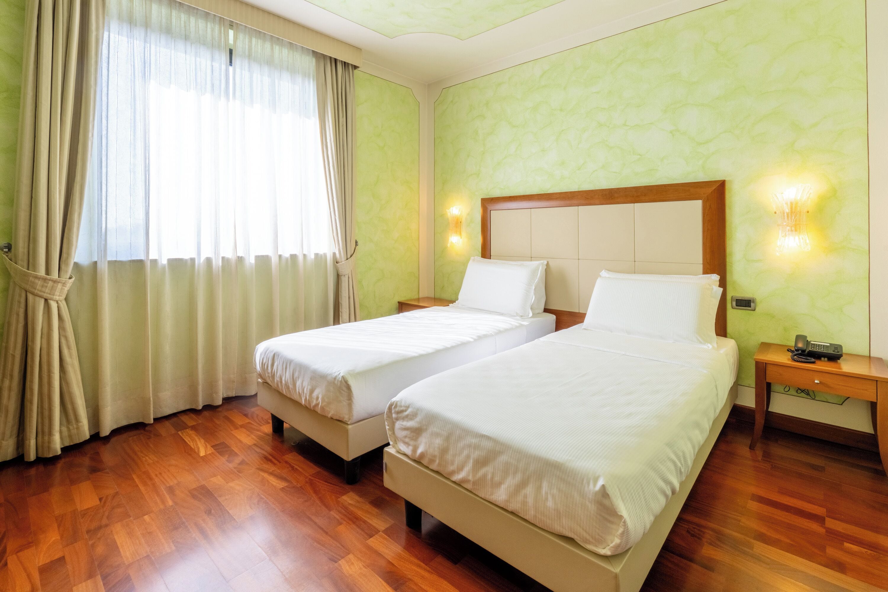 A hotel room with two beds, wooden flooring, green walls, and a window with curtains.