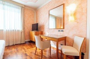 Una camera d'albergo con pavimento in legno, una scrivania con due sedie, un televisore, uno specchio e un lampadario.