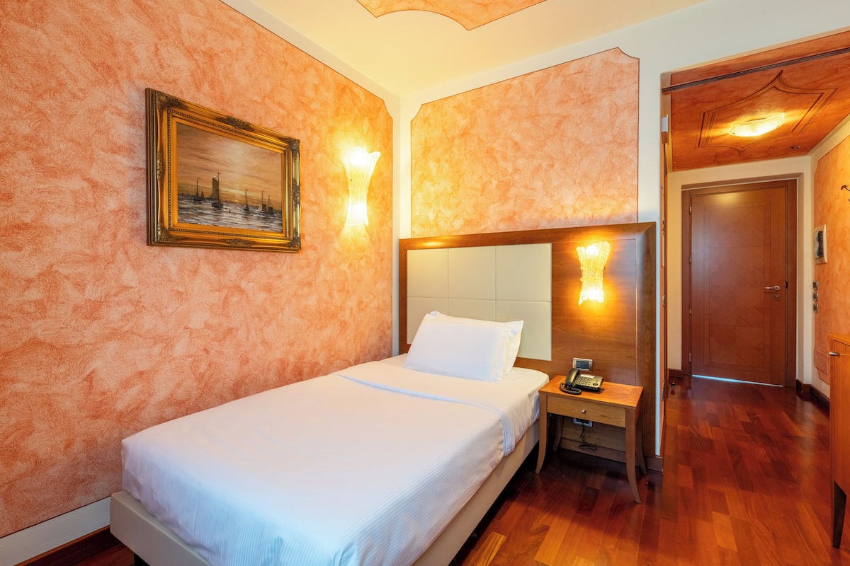 A hotel room with a bed, a nightstand with a phone, a painting on the wall, and a wooden door.