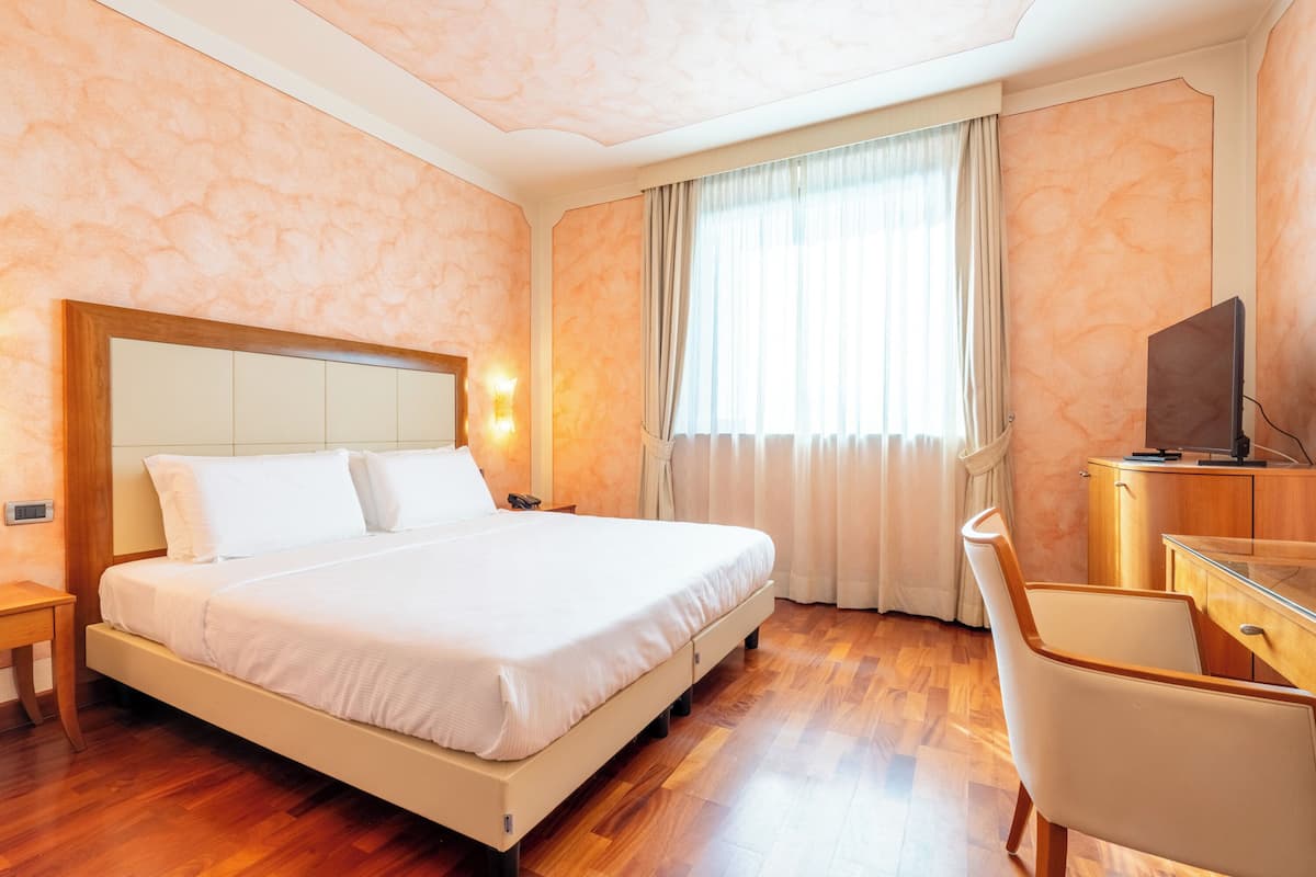 A hotel room with a large bed, a desk, a chair, a television, and a window with curtains.