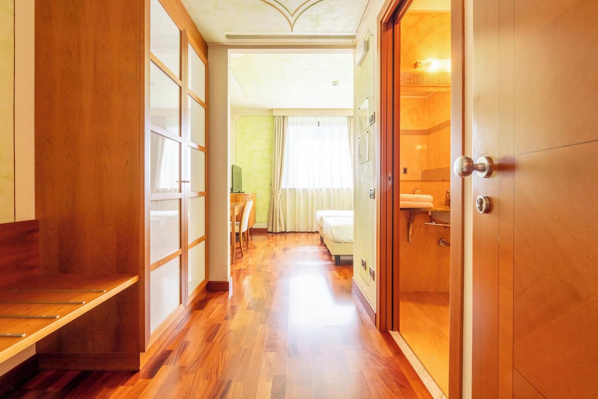 A hotel corridor with wooden flooring, a bed, a desk, and a wardrobe.