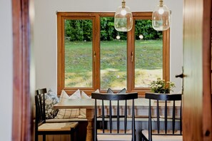 Ein Essbereich mit Holztisch und -stühlen, einer Bank mit Kissen und Blick durch große Fenster auf eine grüne Außenanlage.