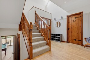 Eine Treppe mit hölzernen Geländern führt zu einer Empore, eine Holztür mit einem runden Spiegel darüber und ein Blick ins Freie durch eine Glastür.