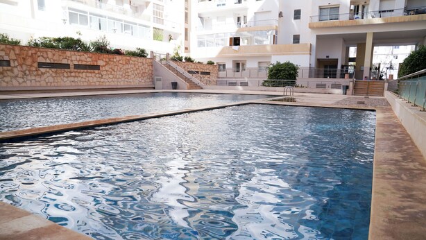 A swimming pool with a stone wall, steps leading to a balcony, and a building with multiple floors in the background. - Chic Designer Apartment with Pool Access (Agadir)