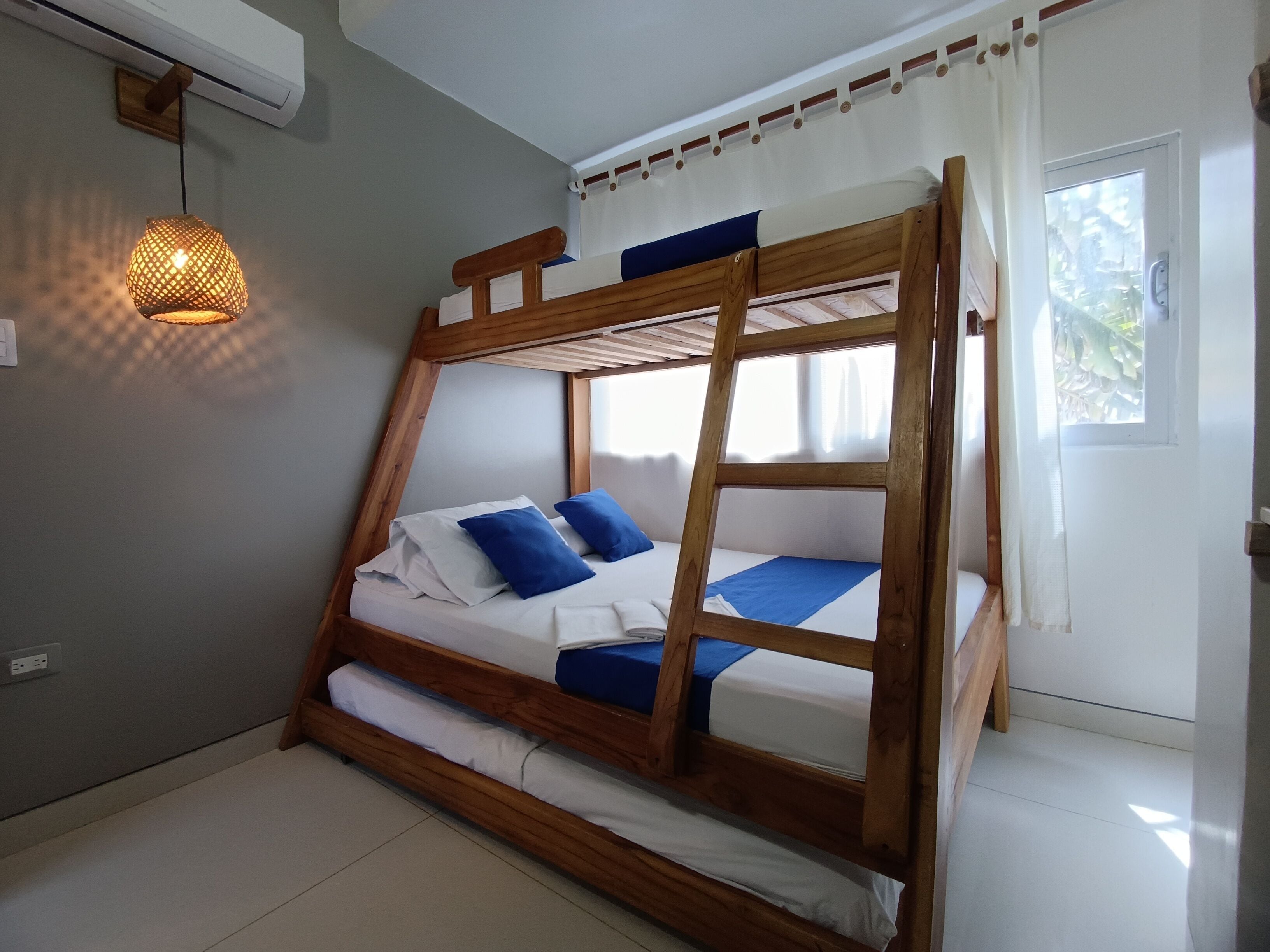 Una cama litera con estructura de madera, ropa de cama blanca y almohadas azules. Hay un colgante de luz y una ventana con cortinas.