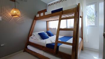 Una cama litera con estructura de madera, ropa de cama blanca y almohadas azules. Hay un colgante de luz y una ventana con cortinas.