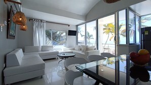A modern living room with a sofa, chairs, a glass table, and large windows offering a view of palm trees and the ocean. - Hotel Portoalegre (Coveñas)