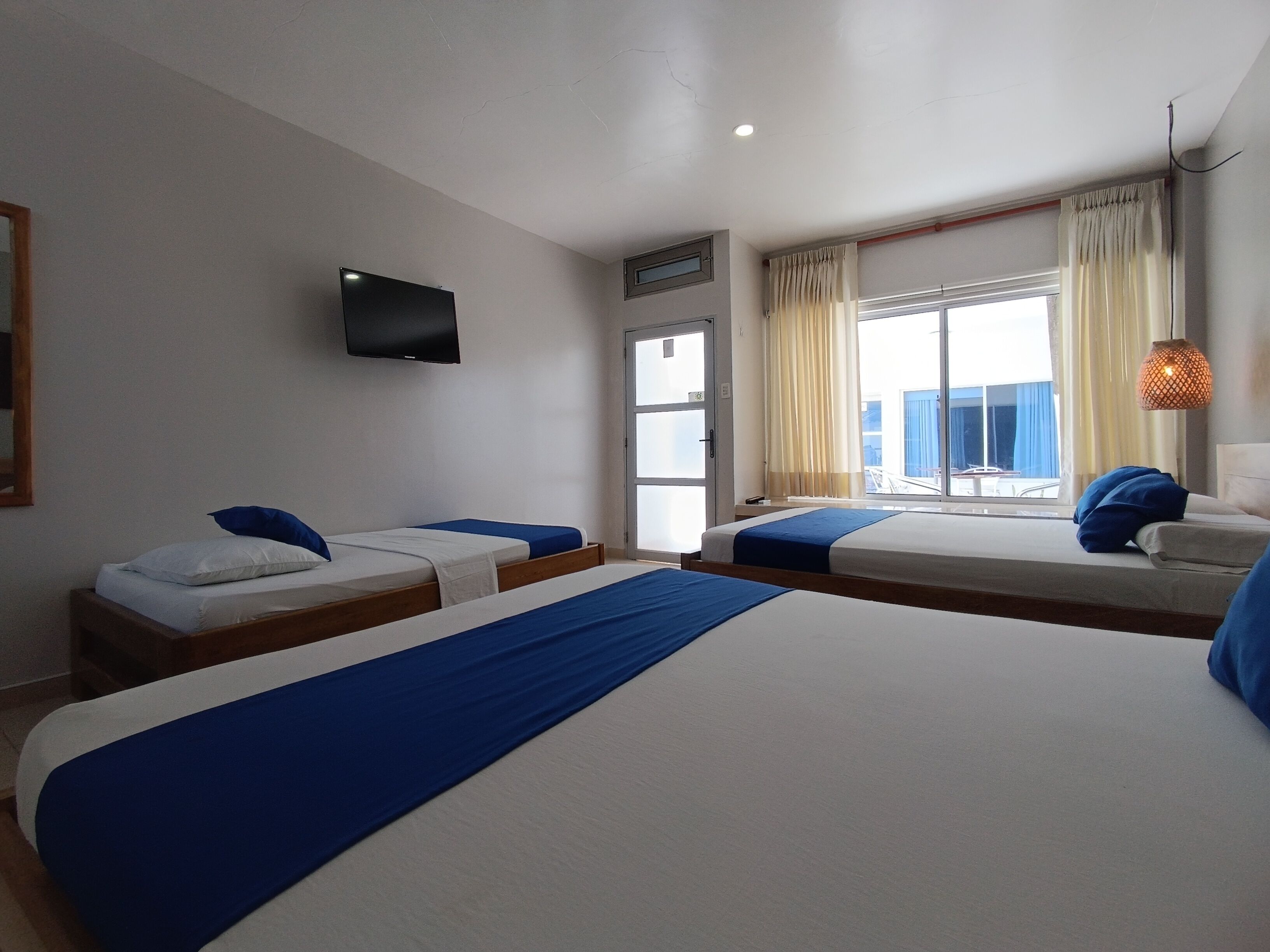 A hotel room with a large bed, two smaller beds, a TV, a window with curtains, and a balcony view.